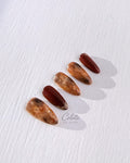 Handmade tortoiseshell press-on nails in translucent amber-brown colour and deep burgundy, finished with gold foil accents on a textured white background.
