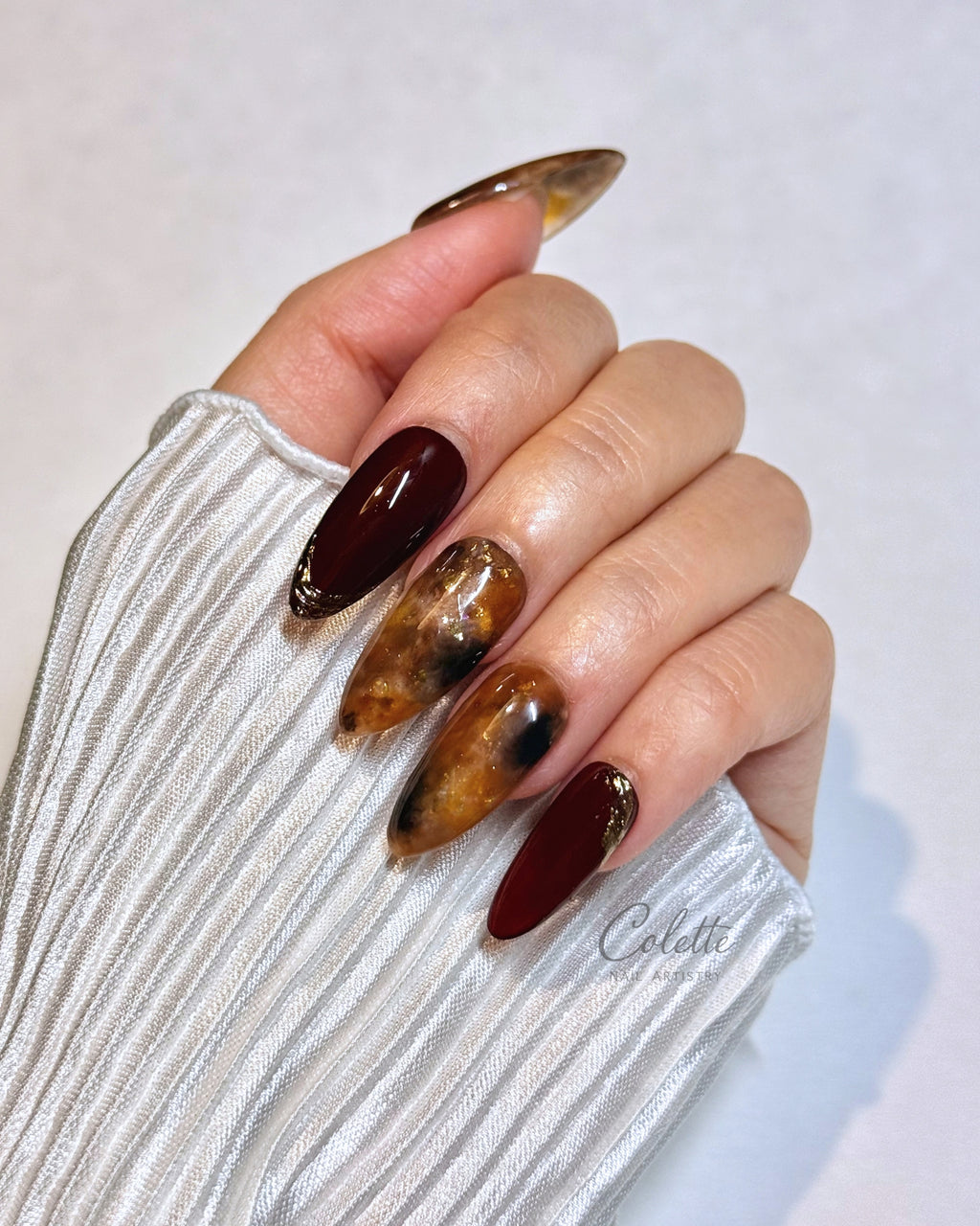 A woman wearing tortoiseshell press-on nails by Colette Nail Artistry, featuring translucent brown marbling, rich burgundy nails and delicate gold foil details.