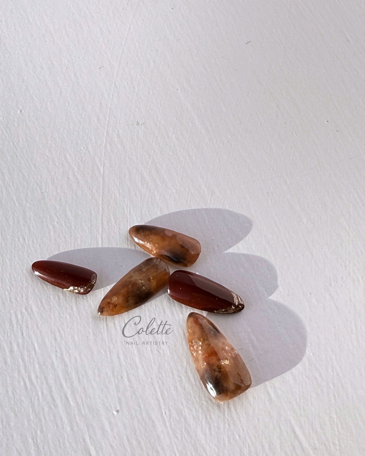 Artistic arrangement of tortoiseshell press-on nails in warm brown and burgundy tones, with translucent marbled layers and gold foil accents in natural sunlight.