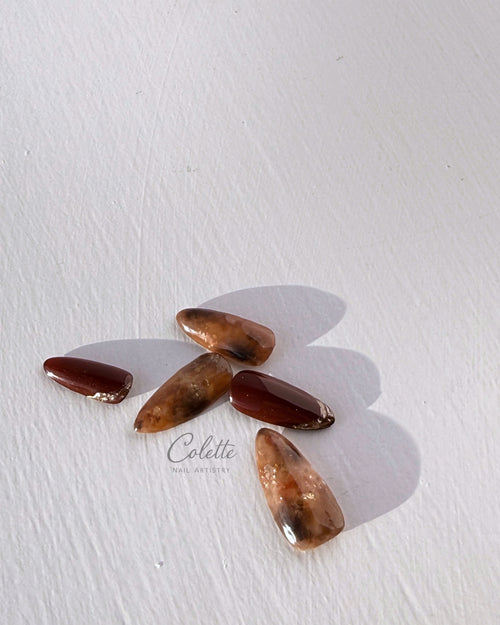 Artistic arrangement of tortoiseshell press-on nails in warm brown and burgundy tones, with translucent marbled layers and gold foil accents in natural sunlight.